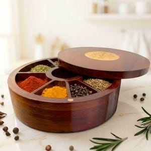 May include: A round wooden spice box with a removable lid featuring a central brass inlay. The container is divided into seven distinct compartments, each filled with various colorful spices including red chili powder, yellow turmeric, and whole cloves.