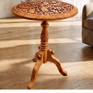 May include: A round wooden side table featuring an intricate floral carving on the tabletop surface. The table stands on a central pedestal base with a carved pineapple-like detail and three curved legs that extend outward for stability.