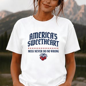 Könnte beinhalten: Ein weißes kurzärmeliges T-Shirt mit dem dunkelblauen Schriftzug AMERICA'S SWEETHEART über einer Reihe roter Sterne und dem blauen Schriftzug MISS NEVER DO NO WRONG sowie einer Grafik aus drei roten Kirschen mit grünen Blättern in der Mitte.