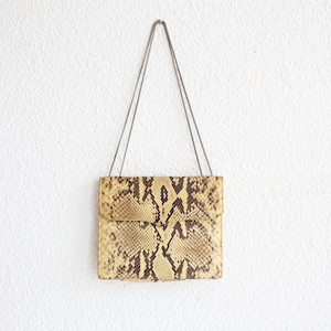 May include: A rectangular handbag with a flap closure, featuring a snakeskin pattern in shades of tan, brown, and cream. The bag hangs from a thin, silver-coloured chain. The background is a plain white wall.