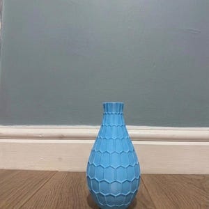 May include: A light blue vase with a textured, repeating hexagonal honeycomb pattern across its exterior. The vase features a narrow neck that flares slightly at the top opening and a rounded, bulbous body that tapers towards the base.