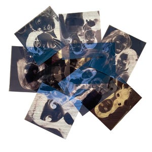 May include: A collection of square photographic prints featuring high-contrast, inverted negative images of various faces and figures. The prints are layered in a scattered arrangement, displaying monochromatic tones with deep shadows and bright highlights.