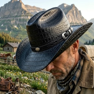 Könnte beinhalten: Schwarzer Cowboyhut mit strukturierter Krokodil-Optik. Der Hut hat eine silberne Schnalle und einen dekorativen Knopf. Der Hut hat eine breite Krempe. Der Hintergrund zeigt Berge und eine rustikale Landschaft.