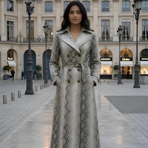 May include: A long, double-breasted trench coat with a snakeskin pattern in shades of gray and white. The coat features a belt at the waist and is worn by a person standing on a cobblestone street.