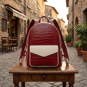 Pode incluir: Uma mochila vermelho-escura com um padrão texturizado de pele de crocodilo, apresentando um bolso frontal contrastante na cor creme com uma aba correspondente, uma alça superior e duas alças ajustáveis com debrum creme nas bordas.