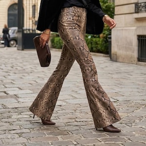 May include: High-waisted trousers featuring a brown and tan snakeskin print pattern. The pants have a flared leg silhouette and are paired with a dark brown clutch bag, a black blazer, and dark brown pointed-toe shoes with a low heel.