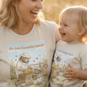 Puede incluir: Camisetas beige con el texto "Un-bee-lievably sweet" y una escena de dibujos animados de un oso apicultor. Una madre y su hijo llevan las camisetas riendo en un campo de flores, bañados por el sol.