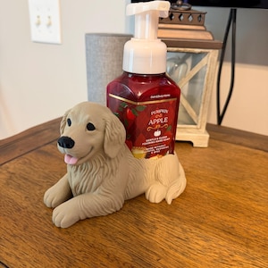 May include: A beige dog-shaped soap holder featuring a sculpted golden retriever lying down, holding a red plastic bottle of Bath & Body Works Pumpkin Apple foaming hand soap with a white pump dispenser and a plaid label design.