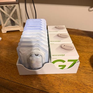 May include: A white plastic organiser tray containing a row of light blue medical sensor applicators and a row of three white boxes labelled Dexcom G7, each featuring a graphic of a round sensor device on the top surface of the packaging.