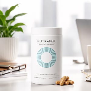 May include: A white cylindrical bottle of Nutrafol Women's Balance hair growth nutraceuticals. The bottle has a teal circular design and text. Several brown capsules are in front of the bottle. A potted plant, laptop, and notebook are in the background.