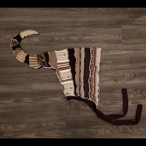 May include: A brown and beige knitted hat with a long tail. The hat is made of different patterned fabrics sewn together. The hat has two long brown ties.