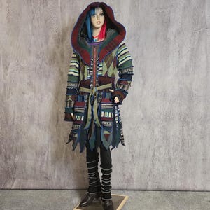 May include: A long, patchwork coat with a hood, in shades of green, blue, red, and brown. The coat has a zip, a belt, and decorative details. The mannequin is wearing black leggings and boots. The coat's design is intricate.