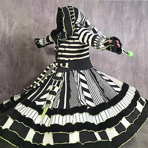 May include: A black and white striped hooded coat with a flared skirt and lime green trim. The garment features a fitted waist with a tie belt, long sleeves with thumb holes, and a hood with a small lime green ball at the tip. The skirt has multiple horizontal ruffles and varied stripe patterns throughout the design.