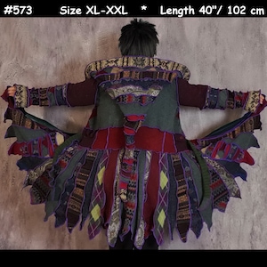 May include: A long, multi-coloured patchwork cardigan with a hood. The cardigan is made of different fabrics and textures, including wool, cotton, and fleece. The cardigan has a fringed hem and is decorated with purple trim. The cardigan is size XL-XXL and is 102 cm long. #573