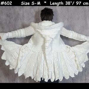 Peut inclure: Un manteau blanc à capuche présentant un design en tricot évasé et texturé avec divers motifs de torsades et des bords volantés. Le vêtement est étiqueté #602, taille S-M, et mesure 97 cm de long. Le manteau affiche une construction complexe.