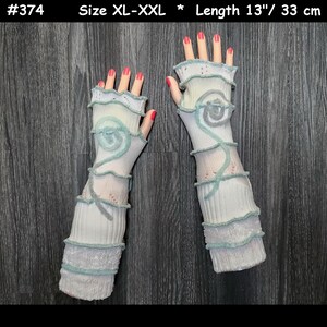 May include: A pair of off-white fingerless gloves with a light blue spiral design and trim. The gloves are approximately 13 inches long and are size XL-XXL. The gloves have a textured appearance and are displayed on a dark wood grain surface.