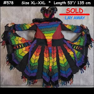 May include: A long coat in size XL-XXL with a length of 135 cm, featuring a vibrant rainbow-coloured patchwork design made of various fabric strips, a black faux fur collar, and a flared hemline with dangling fabric fringe accents.
