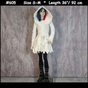 May include: A cream-coloured hooded coat with a textured, layered knit design, long sleeves, and a tie-waist closure. The garment is labelled #605, size S-M, and measures 92 cm in length. It is worn over black leggings and black leg warmers.