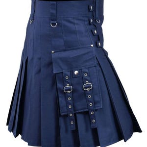 Op de afbeelding: Een marineblauwe utility kilt met een geplooid ontwerp, een grote cargozak aan de voorzijde met zilverkleurige drukknopen en twee verticale banden met metalen gespen en oogjes. De tailleband is voorzien van riemlussen en zijverstelbanden.