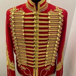 Può includere: Una giacca rossa in stile militare con finiture dorate e dettagli decorativi in corda dorata. La giacca presenta un colletto alto, bottoni dorati e ricami ornati sulle maniche. La giacca è esposta su un manichino nero.