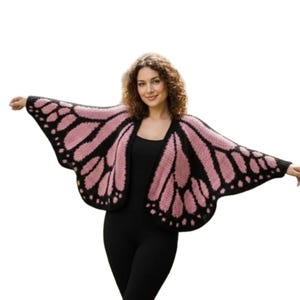 May include: A knitted butterfly wing wrap in black and pink. The wrap is open in the front and has a wing-like design with black outlines and pink filling. The person in the image is wearing a black outfit.