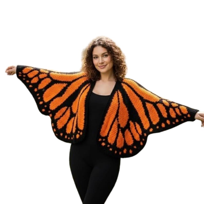 May include: A knitted butterfly wing-shaped cardigan in orange and black. The open-front sweater features a monarch butterfly wing design, with orange sections outlined in black and dotted details. The garment is worn over a black outfit.