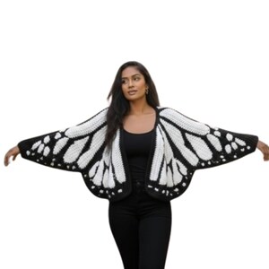May include: A black and white crochet butterfly wing cardigan. The open-front sweater features a black border and wing pattern with white detailing. The model is wearing a black top and black pants.