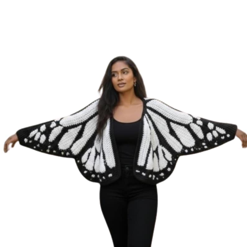 May include: A black and white crochet butterfly wing cardigan. The open-front sweater features a black border and wing pattern with white detailing. The model is wearing a black top and black pants.