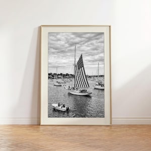 May include: A black and white framed wall art print depicting a sailing boat with a large American flag sail navigating a harbour filled with various other boats under a cloudy sky. A small motorboat with two people is visible in the foreground.