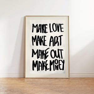 May include: A framed art print featuring the words "MAKE LOVE MAKE ART MAKE OUT MAKE MONEY" in black, bold lettering. The print has a white background and is encased in a gold-coloured frame. It is displayed on a wooden floor.