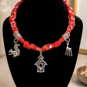 May include: A red fabric necklace with white polka dots and silver charms. The charms include a squirrel, a birdhouse, and a deer. The necklace is displayed on a black mannequin.
