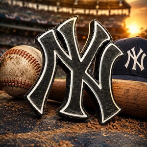 Op de afbeelding: Een zwart-wit New York Yankees logo-embleem geplaatst voor een versleten honkbal, een houten honkbalknuppel en een marineblauwe honkbalpet met hetzelfde witte logo op de voorkant, tegen een wazige achtergrond van een stadion.