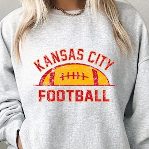 Może przedstawiać: Jasnoszara bluza z okrągłym dekoltem z postarzaną czerwoną i ż&oacute;łtą grafiką na klatce piersiowej. Wz&oacute;r zawiera napis KANSAS CITY wygięty w łuk nad ż&oacute;łtą ilustracją piłki do futbolu amerykańskiego z czerwonymi akcentami oraz słowo FOOTBALL poniżej.