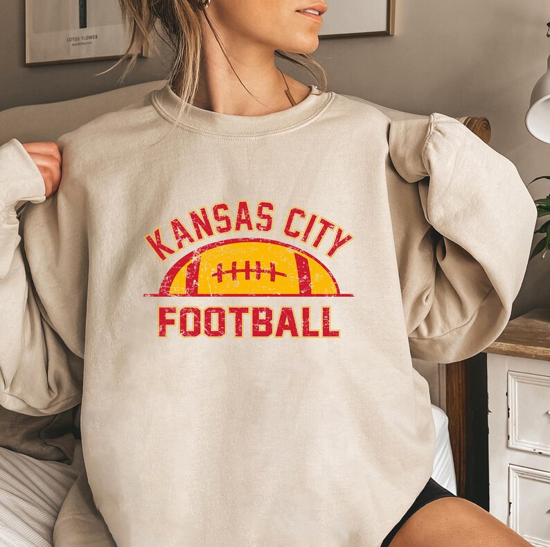 Może przedstawiać: Beżowa bluza z okrągłym dekoltem i nadrukiem graficznym w kolorze czerwonym i ż&oacute;łtym z efektem postarzenia. Wz&oacute;r zawiera napis KANSAS CITY czerwoną, łukowatą czcionką nad ż&oacute;łtą ilustracją piłki do futbolu amerykańskiego oraz słowo FOOTBALL.