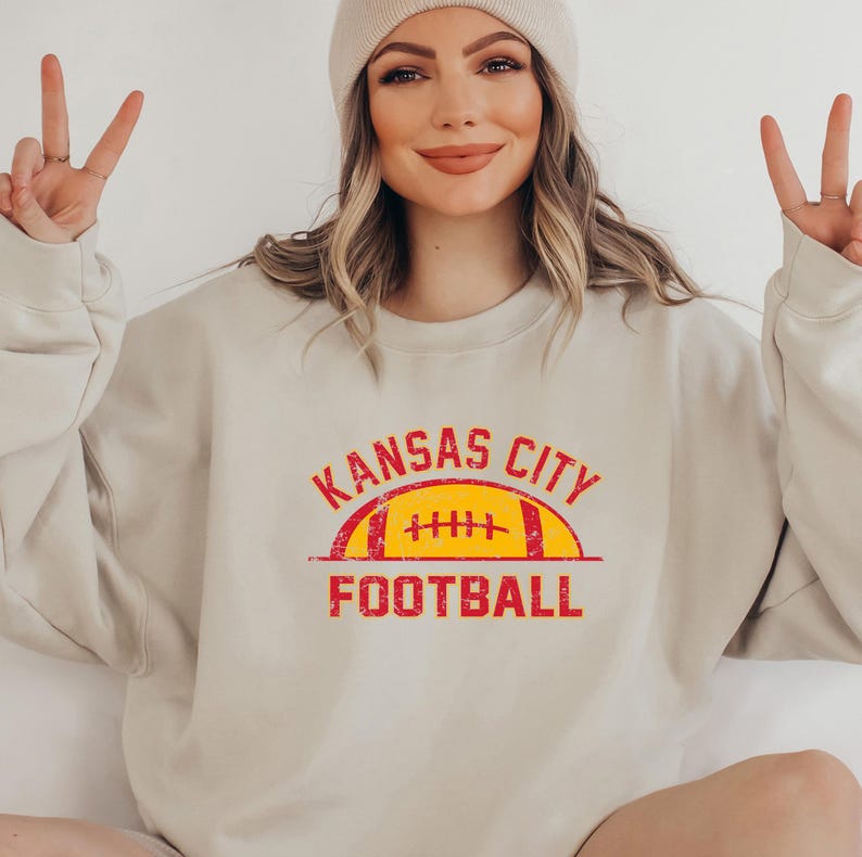 Może przedstawiać: Beżowa bluza z okrągłym dekoltem i postarzaną czerwono-ż&oacute;łtą grafiką na klatce piersiowej. Wz&oacute;r zawiera napis KANSAS CITY wygięty w łuk nad ż&oacute;łtą ilustracją piłki do futbolu amerykańskiego oraz słowo FOOTBALL nadrukowane na czerwono pod grafiką.