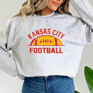 Może przedstawiać: Jasnoszara bluza z okrągłym dekoltem i postarzanym nadrukiem w kolorze czerwonym i ż&oacute;łtym. Wz&oacute;r zawiera napis KANSAS CITY w łuku nad ilustracją ż&oacute;łtej piłki do futbolu amerykańskiego z czerwonymi sznurowadłami oraz słowo FOOTBALL poniżej.