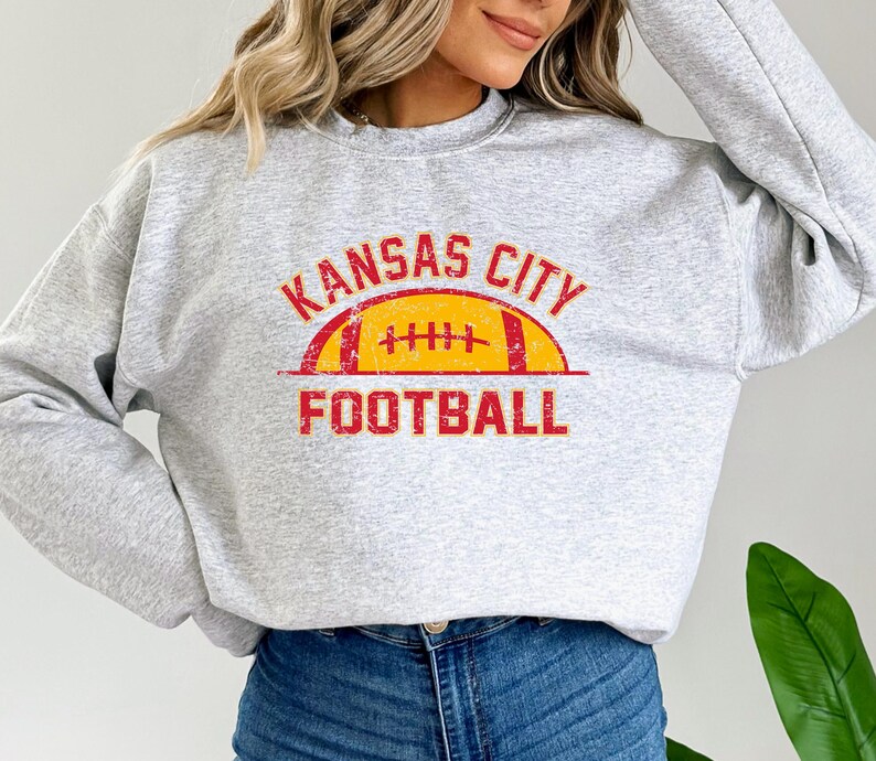 Może przedstawiać: Jasnoszara bluza z okrągłym dekoltem i postarzanym nadrukiem w kolorze czerwonym i ż&oacute;łtym. Wz&oacute;r zawiera napis KANSAS CITY w łuku nad ilustracją ż&oacute;łtej piłki do futbolu amerykańskiego z czerwonymi sznurowadłami oraz słowo FOOTBALL poniżej.