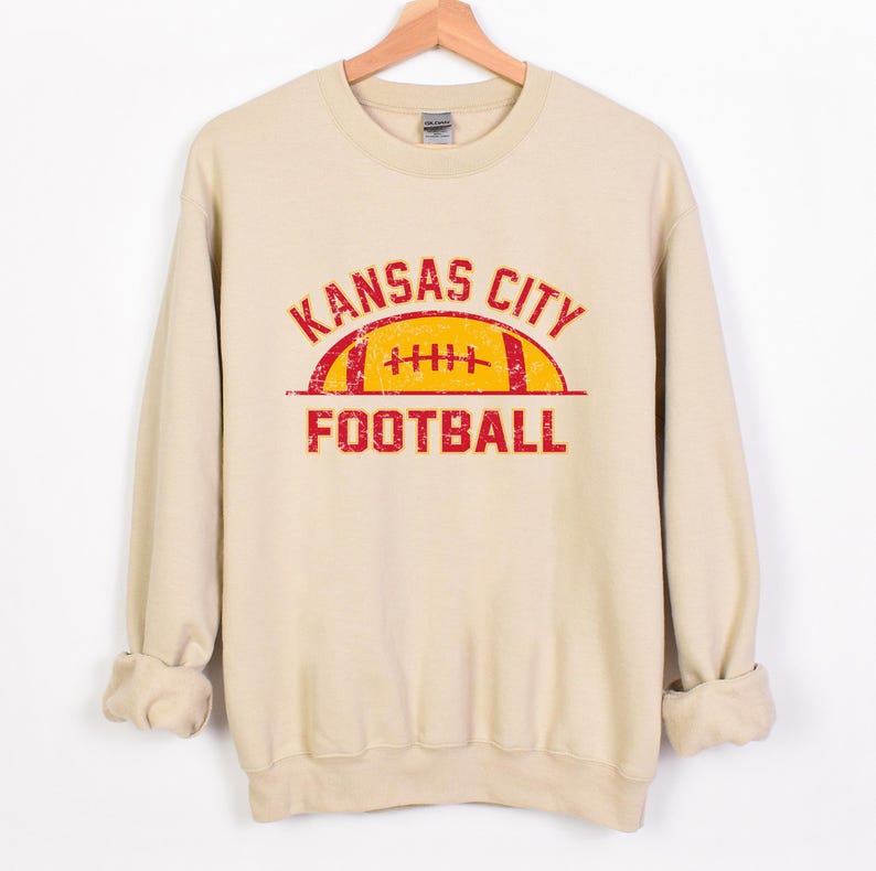 Może przedstawiać: Beżowa bluza z okrągłym dekoltem i postarzanym czerwonym nadrukiem z napisem KANSAS CITY FOOTBALL w łukowatym kroju nad stylizowaną ilustracją ż&oacute;łto-czerwonej piłki do futbolu amerykańskiego. Odzież posiada długie rękawy oraz ściągacze przy mankietach.