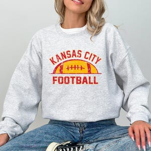 Może przedstawiać: Jasnoszara bluza z okrągłym dekoltem z postarzanym czerwono-ż&oacute;łtym nadrukiem na klatce piersiowej. Wz&oacute;r zawiera napis KANSAS CITY wygięty w łuk nad ilustracją ż&oacute;łtej piłki do futbolu z czerwonymi sznurowadłami oraz słowo FOOTBALL poniżej.