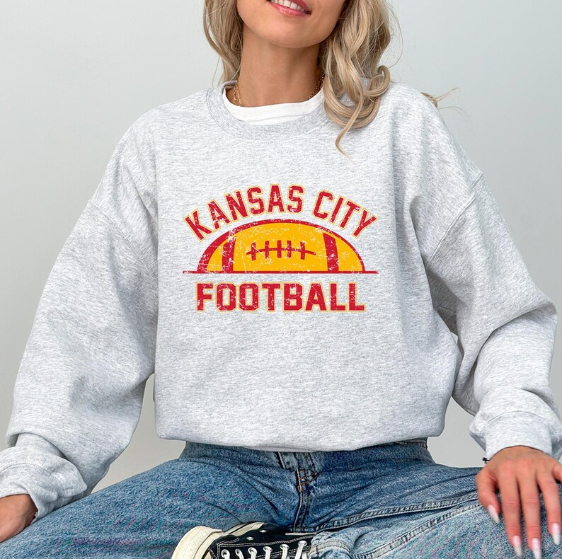Może przedstawiać: Jasnoszara bluza z okrągłym dekoltem z postarzanym czerwono-ż&oacute;łtym nadrukiem na klatce piersiowej. Wz&oacute;r zawiera napis KANSAS CITY wygięty w łuk nad ilustracją ż&oacute;łtej piłki do futbolu z czerwonymi sznurowadłami oraz słowo FOOTBALL poniżej.