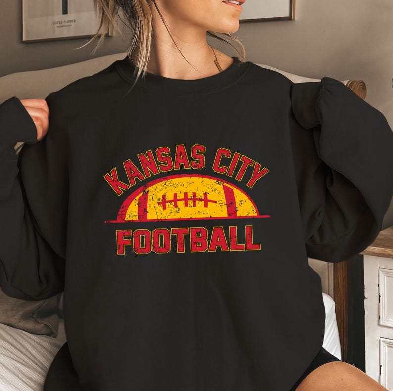 Może przedstawiać: Czarna bluza z okrągłym dekoltem z postarzanym nadrukiem graficznym w kolorze czerwonym i ż&oacute;łtym. Wz&oacute;r zawiera napis KANSAS CITY wygięty w łuk nad stylizowaną ilustracją piłki do futbolu oraz słowo FOOTBALL wydrukowane pogrubioną czcionką.