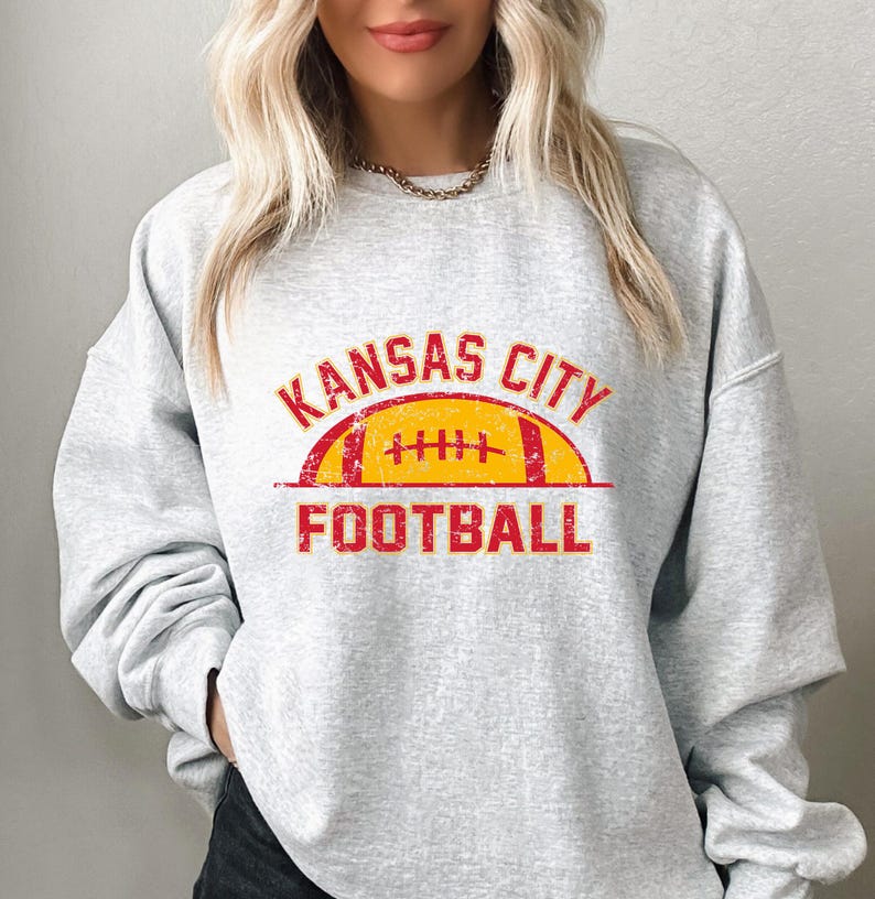 Może przedstawiać: Jasnoszara bluza z okrągłym dekoltem z postarzaną czerwoną i ż&oacute;łtą grafiką na klatce piersiowej. Wz&oacute;r zawiera napis KANSAS CITY wygięty w łuk nad ż&oacute;łtą ilustracją piłki do futbolu amerykańskiego z czerwonymi akcentami oraz słowo FOOTBALL poniżej.