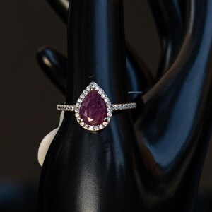 May include: A pear-shaped ruby ring featuring a halo of small white diamonds set in a thin metallic band. The central deep red gemstone is surrounded by a delicate border of sparkling stones, creating a classic and elegant jewelry piece.