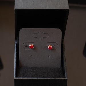 May include: A pair of round, faceted red stud earrings secured to a black rectangular display card. The earrings are presented inside an open, black textured jewelry box. The text Aliona Lagoda is visible in the bottom left corner of the image.