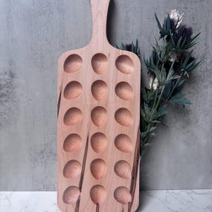 May include: A rectangular light-colored wooden serving board featuring a handle with a circular hole at the top. The board contains eighteen evenly spaced, rounded indentations arranged in three vertical columns of six, with visible wood grain.