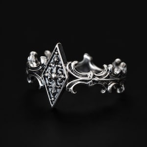 Puede incluir: Un anillo de plata con un diseño ornamentado. El anillo presenta un elemento central en forma de diamante con un patrón floral detallado y un pequeño acento circular. La banda tiene un diseño decorativo y fluido.