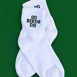 May include: A pair of white crew socks featuring ribbed cuffs and the text GO BIRDS DH embroidered in dark green thread on the upper portion of each sock. The socks are positioned overlapping each other, showcasing the clean white fabric.