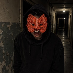 May include: A black hooded sweatshirt worn with a textured, orange, fungus-like mask covering the upper face. The mask features intricate, brain-like folds and ridges, while the mouth area shows visible teeth and dark red, blood-like stains.