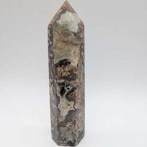 Puede incluir: Una torre de piedra pulida con base hexagonal y punta. La superficie presenta una mezcla de tonos tierra marrones, beige y gris translúcido con una prominente inclusión cavernosa de textura rugosa que recorre verticalmente la cara frontal.