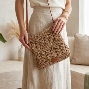 Pode incluir: Bolsa transversal tecida em cor bege natural com um design geométrico de tecido aberto. Possui uma alça de couro marrom e uma pequena etiqueta de couro. A bolsa é segurada por uma pessoa vestindo uma roupa em tons neutros.